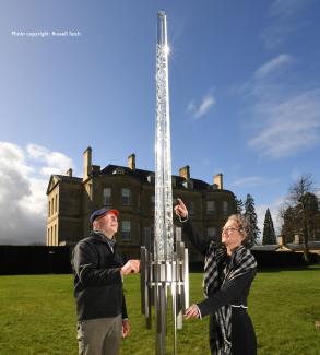 two people smiling looking up at monumental glass spire sculpture on lawn in front of large english country house with blue sky